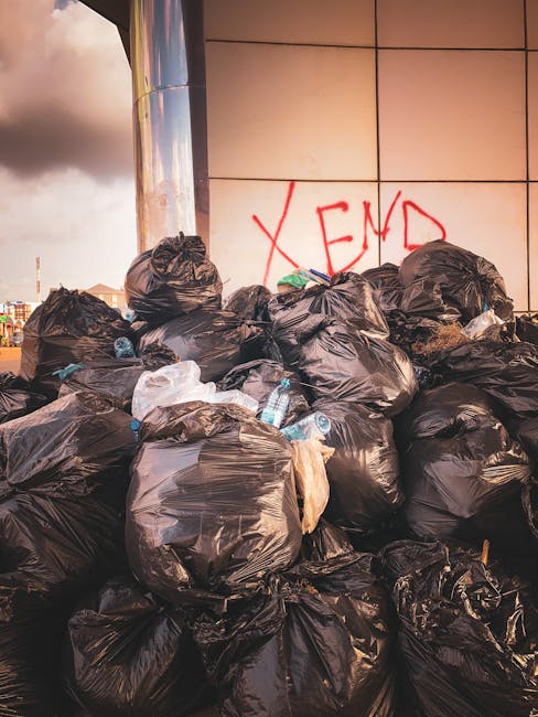 A collection of overflowing waste and rubbish bags accumulated around multiple recycling bins in an urban outdoor setting. The waste includes black and green plastic trash bags, cardboard boxes, crump